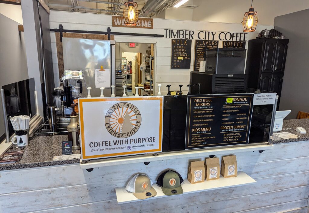 The interior of Timber City Coffee Co. in Plainfield, Indiana, showcasing a welcoming coffee shop atmosphere. There are signs for various coffee drinks, including specialty blends, and a display of baked goods. The shop features a rustic and modern decor with a wooden counter and a "Faith-Family-Friends-Coffee" logo.