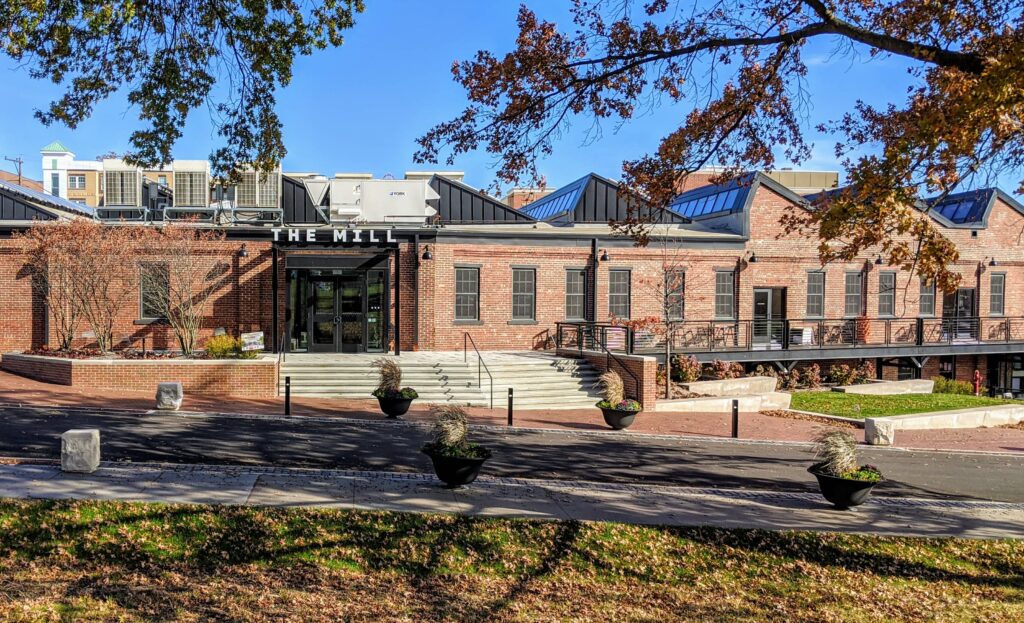 Exterior view of The Mill, a modern co-working space in Bloomington with brick walls, large windows, and a welcoming entrance, surrounded by landscaped greenery and seating areas.