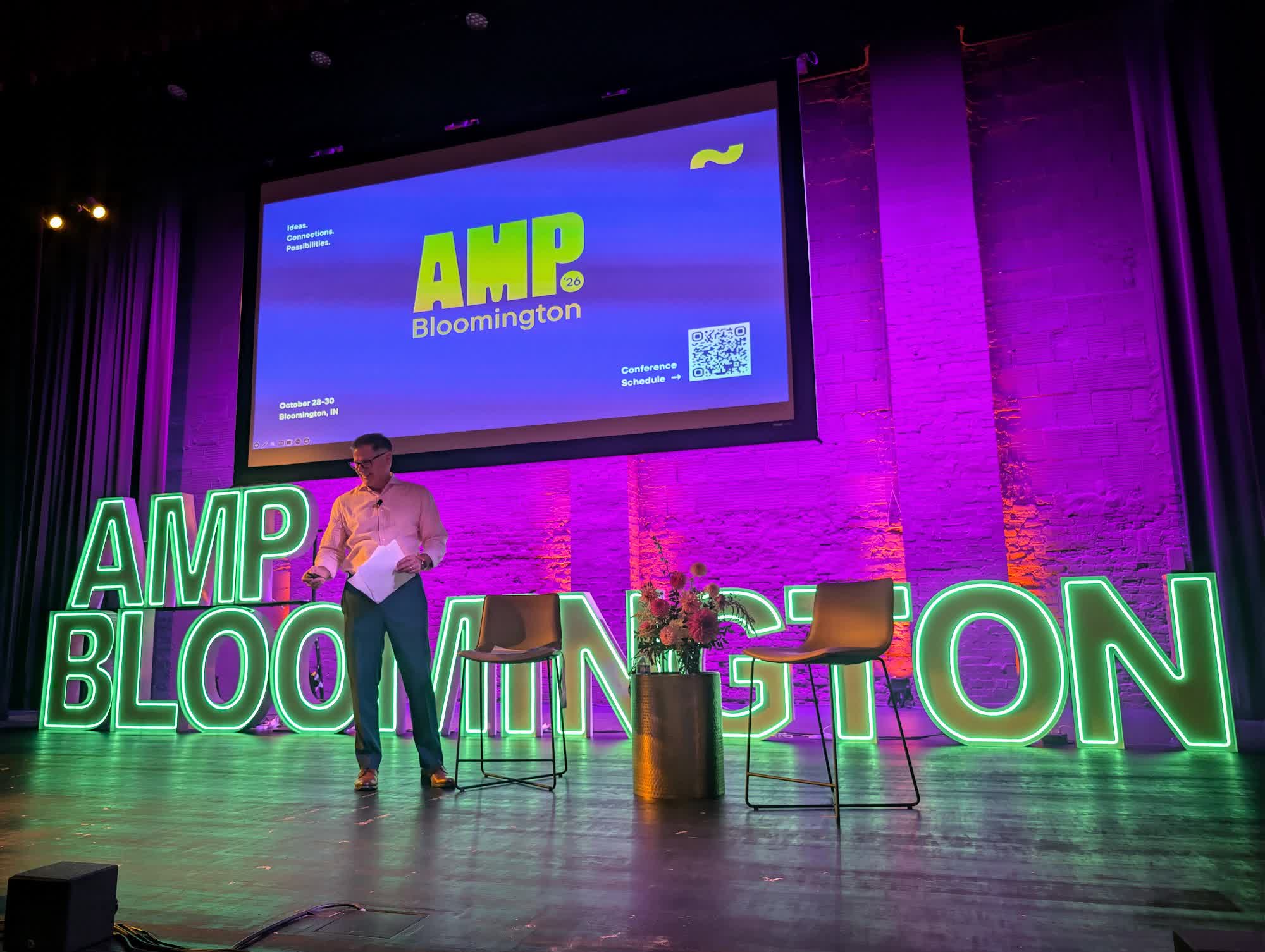 A speaker opens the program on the Buskirk-Chumley stage in front of glowing AMP Bloomington letters and the AMP 26 title slide
