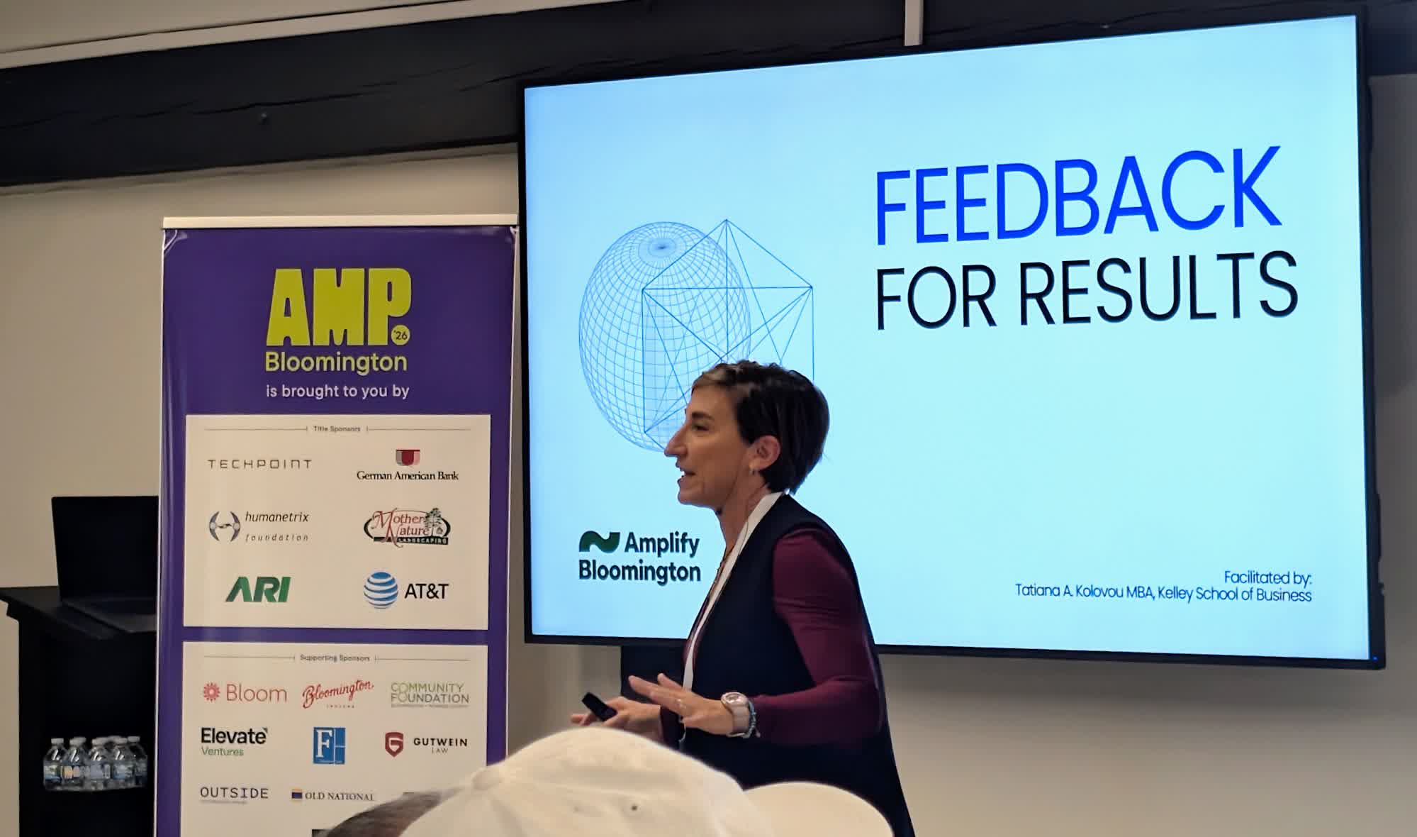 A facilitator presents in front of a slide titled Feedback for Results, with the AMP Bloomington sponsor banner beside the speaker