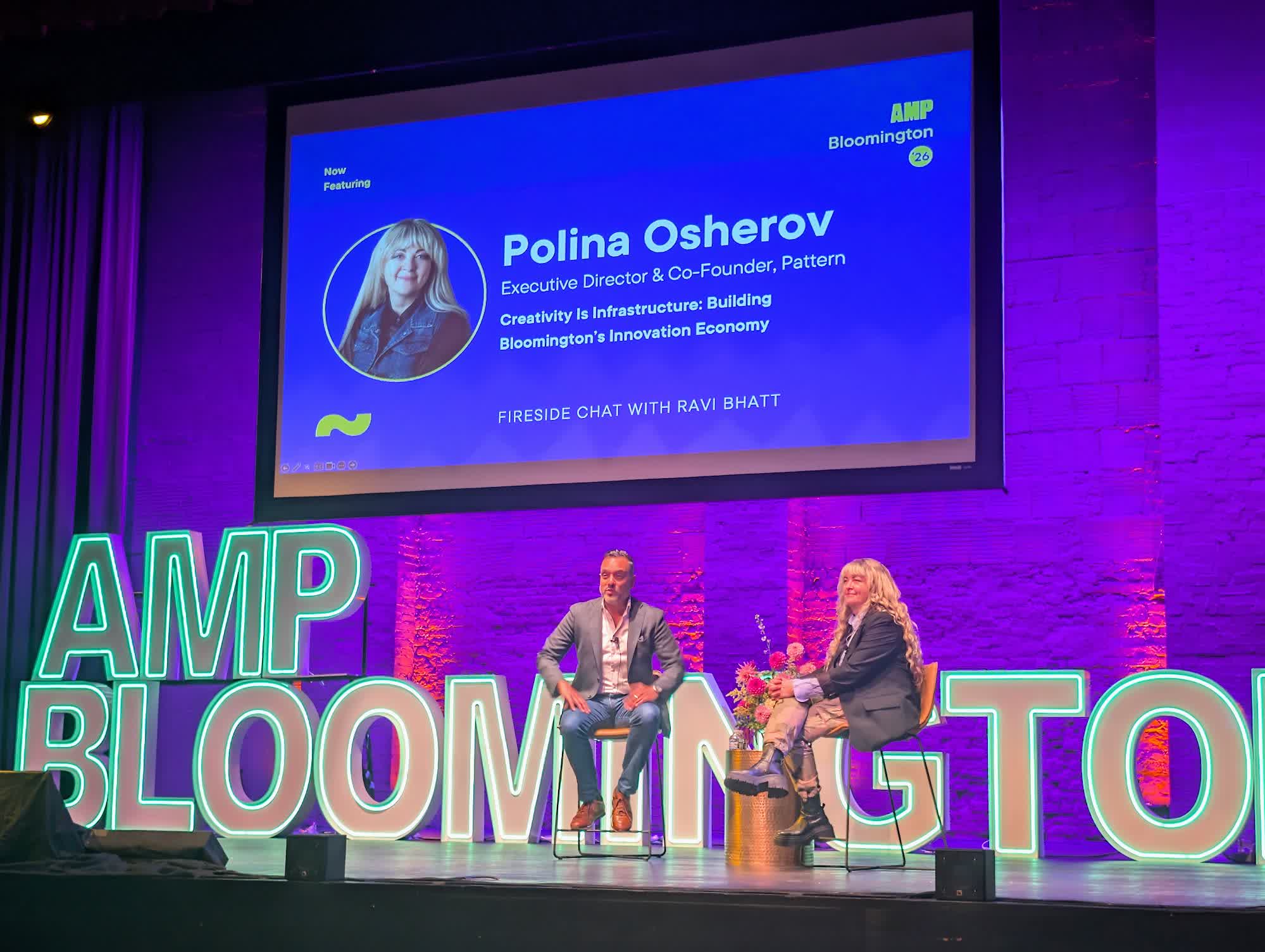 Two people seated on the AMP Bloomington stage for a fireside chat, with a slide introducing Polina Osherov of Pattern and the talk Creativity Is Infrastructure