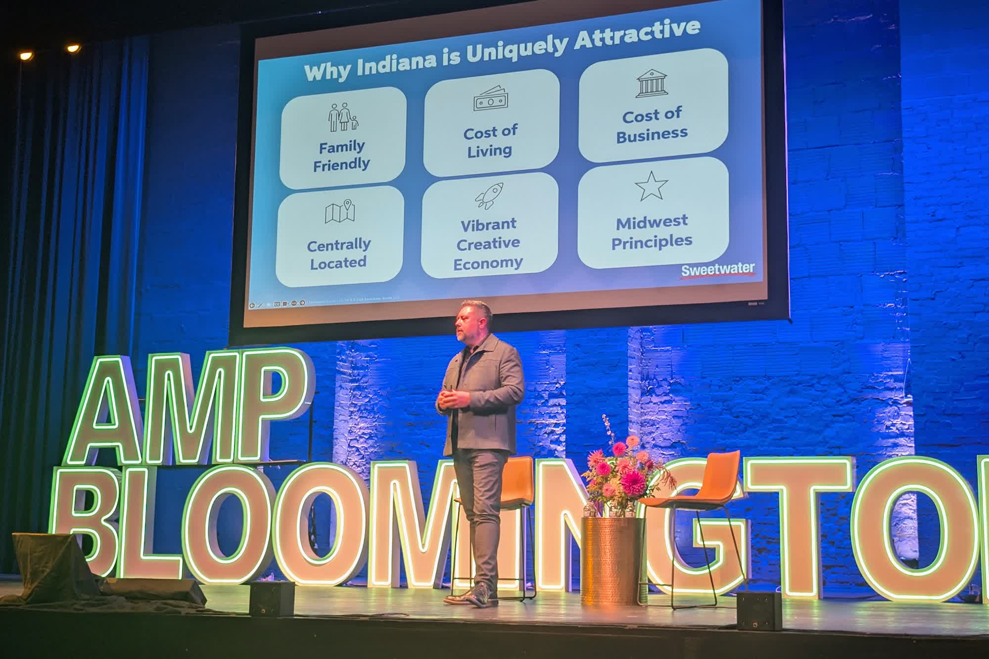 A speaker on the AMP Bloomington stage in front of a slide titled Why Indiana is Uniquely Attractive, listing family friendly, cost of living, cost of business, centrally located, vibrant creative economy and Midwest principles