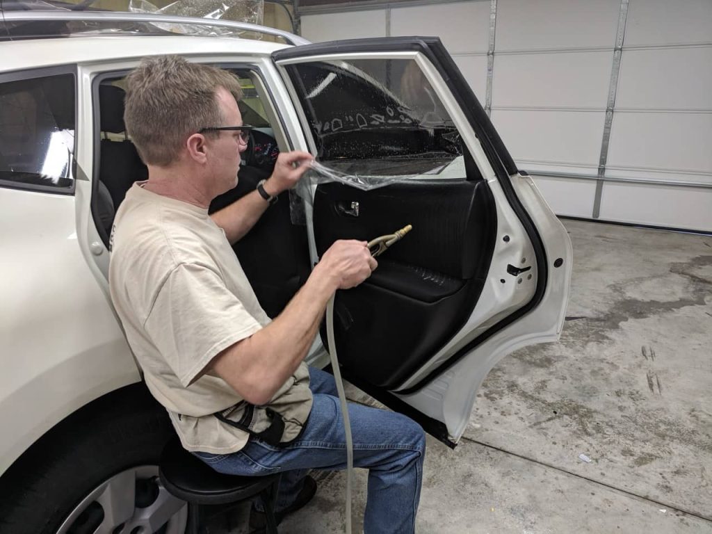 Technician installing window tint film on a car door window using a heat tool inside a garage workshop