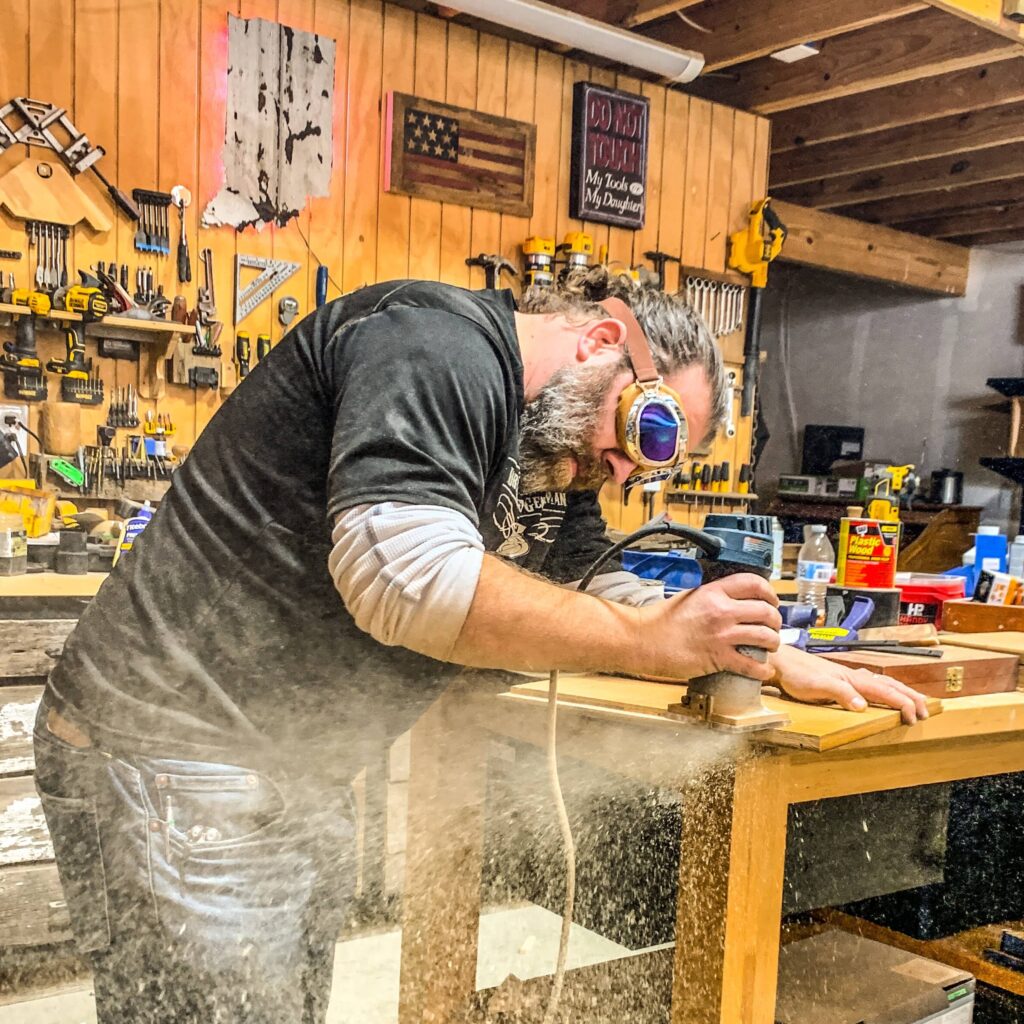 Bobby Williams working on a wood panel with a handheld power tool at a workshop bench.