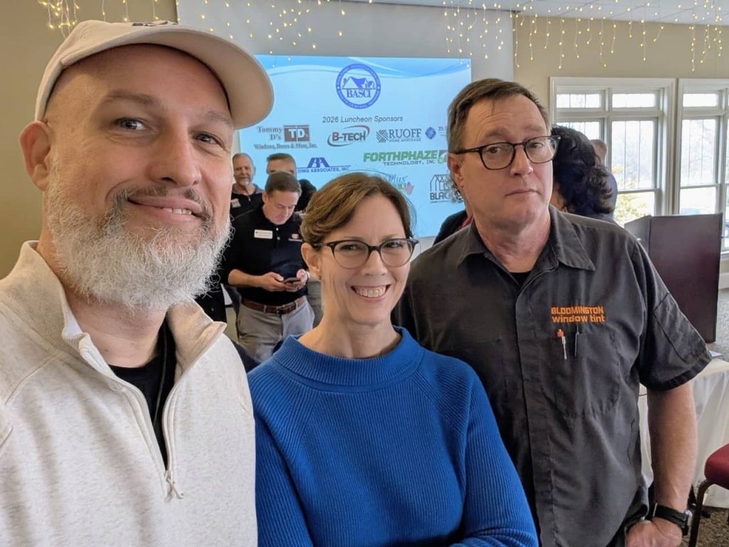 Three attendees posing for a selfie at a Bloomington business networking luncheon with sponsor signage in the background