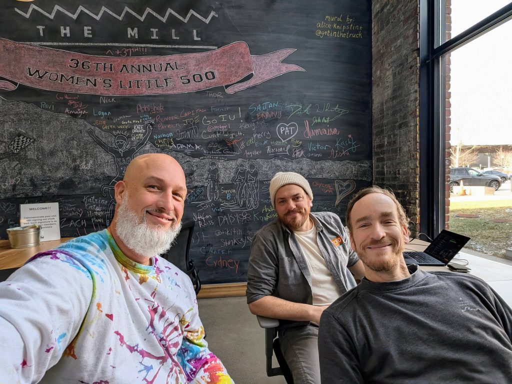 Three men smiling at The Mill coworking space in Bloomington, Indiana, seated in front of a large chalkboard mural referencing the 36th Annual Women's Little 500. Mural by Alice Knipstine.