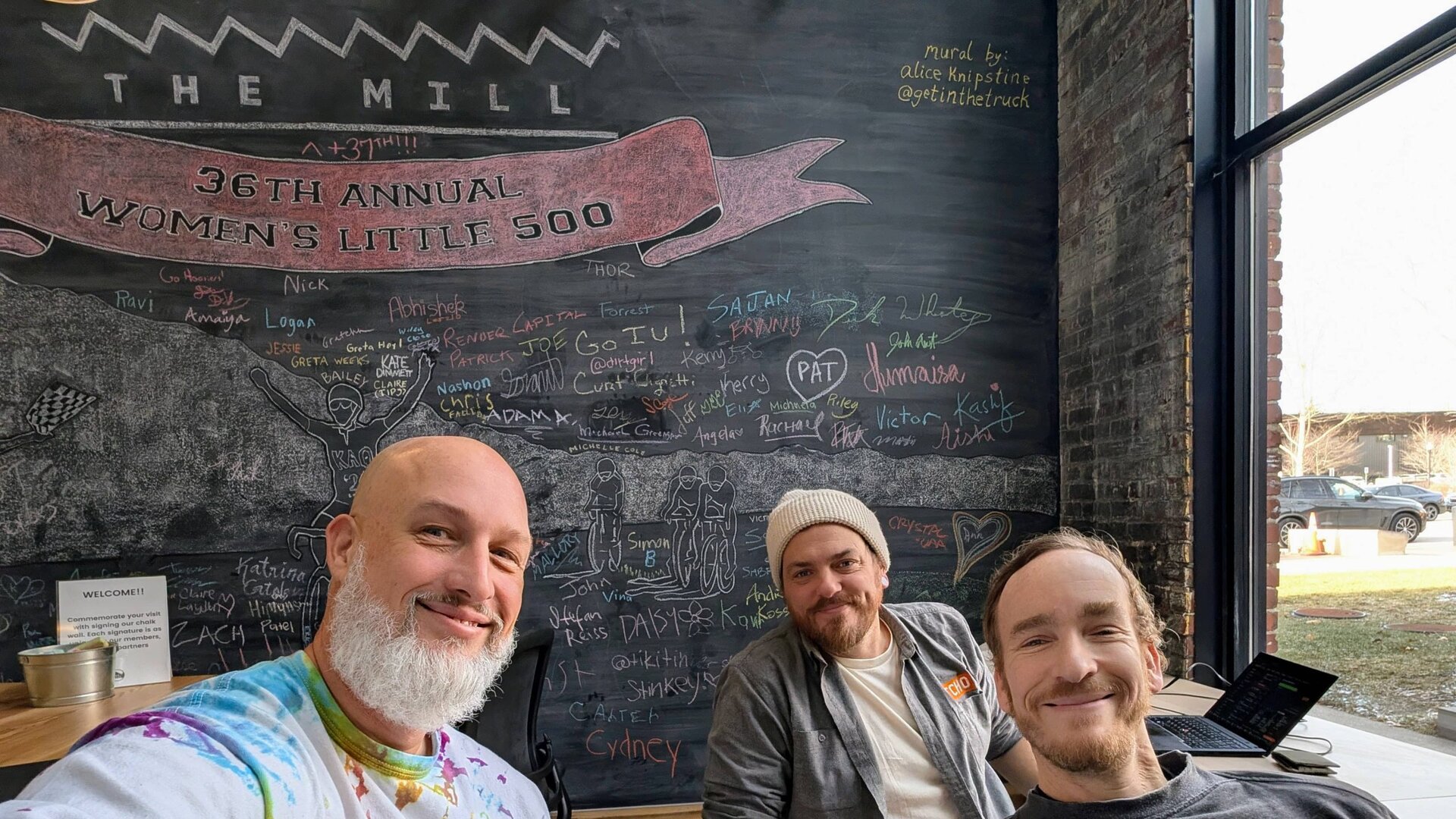 Dave Martin with two people at The Mill in Bloomington, seated in front of a chalkboard mural for the 36th Annual Women's Little 500.