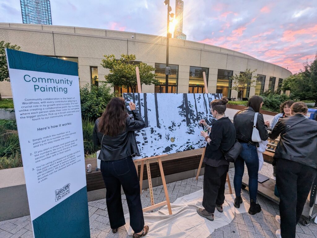 Attendees of WordCamp US 2024 participate in a community painting activity outdoors at the Thank You Social, with a paint-by-numbers mural in progress.