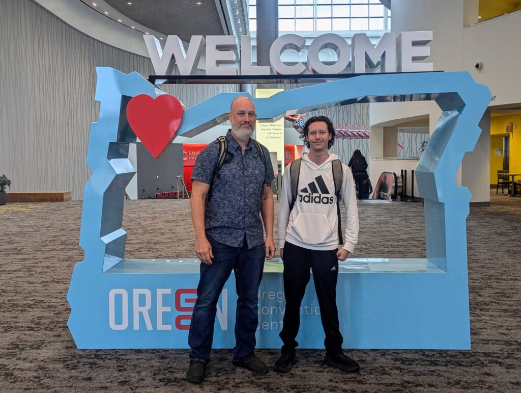 Dave Martin and Jarod Maxwell standing together at the Oregon Convention Center, posing in front of a large "Welcome to Oregon" display with a heart symbol.