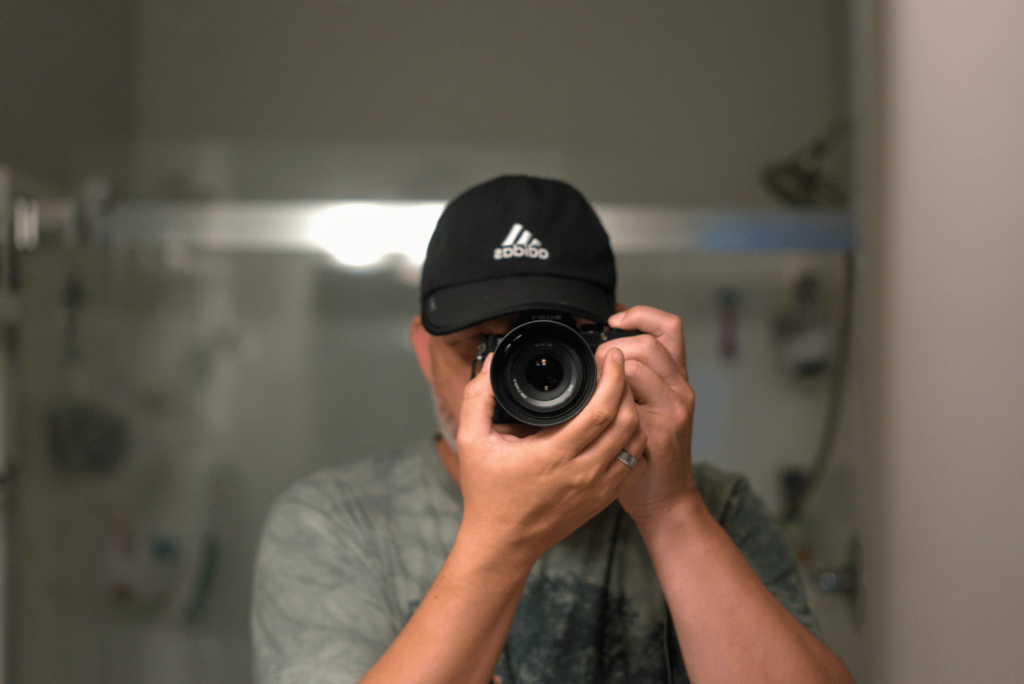 Self-portrait of photographer Dave Martin from David Martin Design, holding a camera up to his face, wearing a black cap, in front of a mirror.