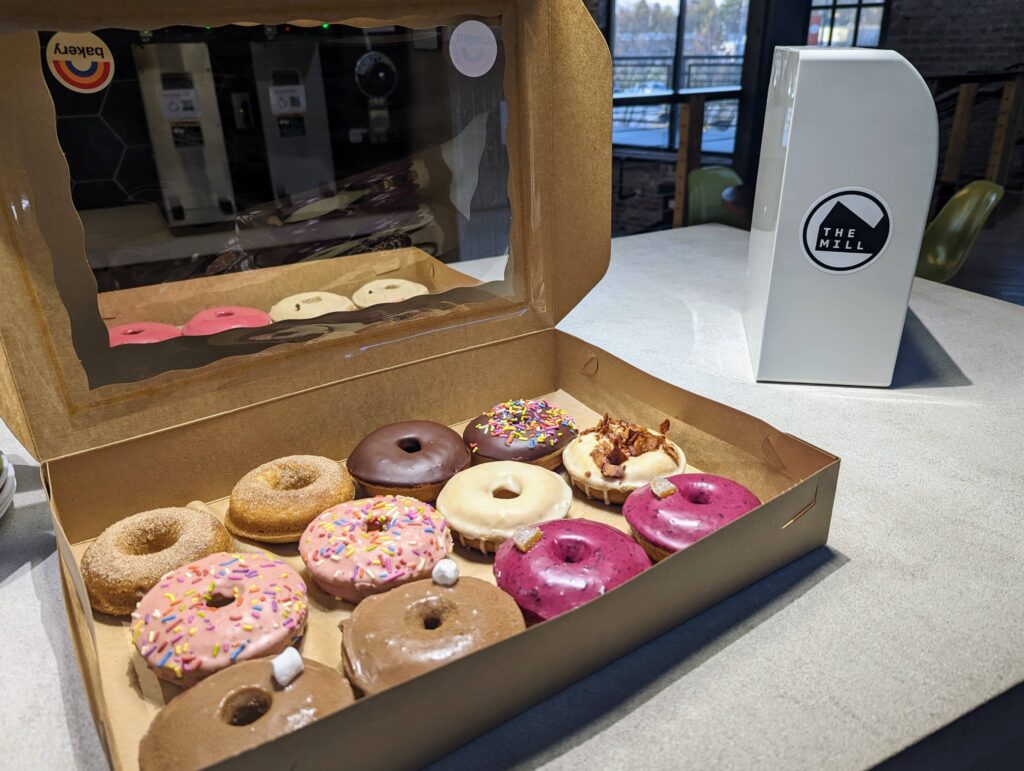 A box of assorted donuts with various toppings, including sprinkles, chocolate, and glaze, set on a table at The Mill in Bloomington, with The Mill’s logo visible in the background.