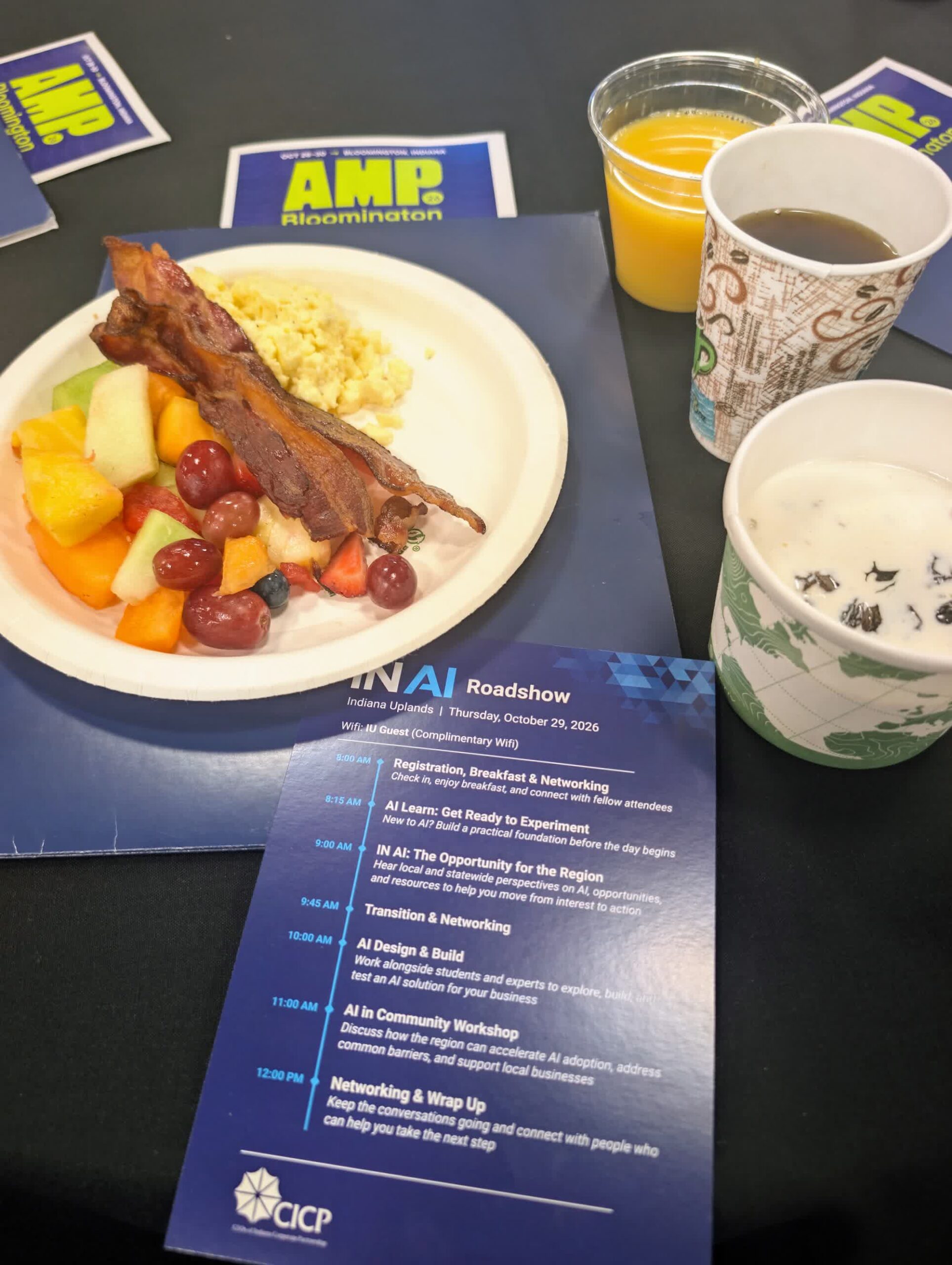 Breakfast plate, coffee and an IN AI Roadshow agenda card on a table with AMP Bloomington stickers