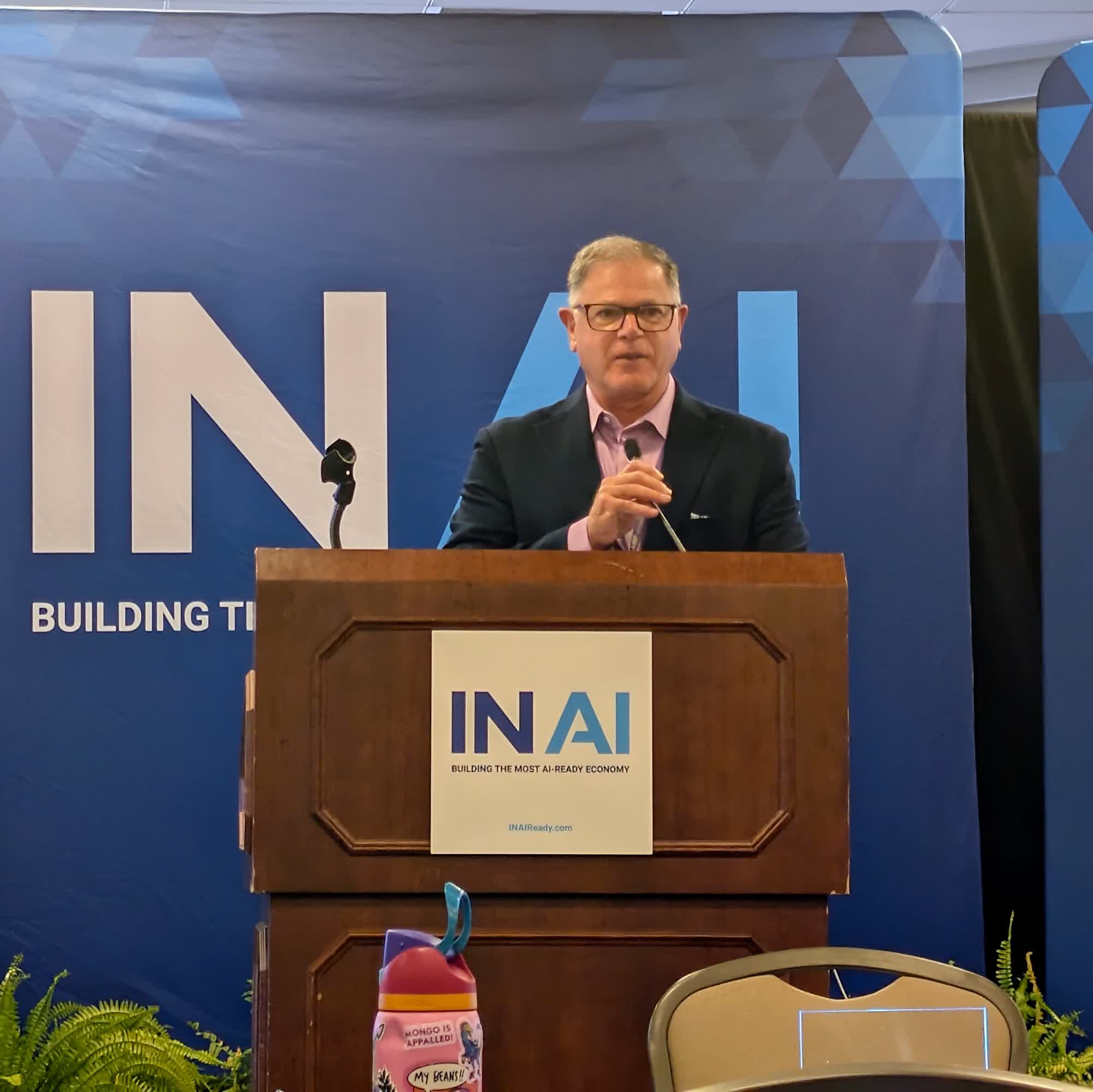 A speaker with glasses stands at a wooden podium marked IN AI in front of a blue IN AI backdrop