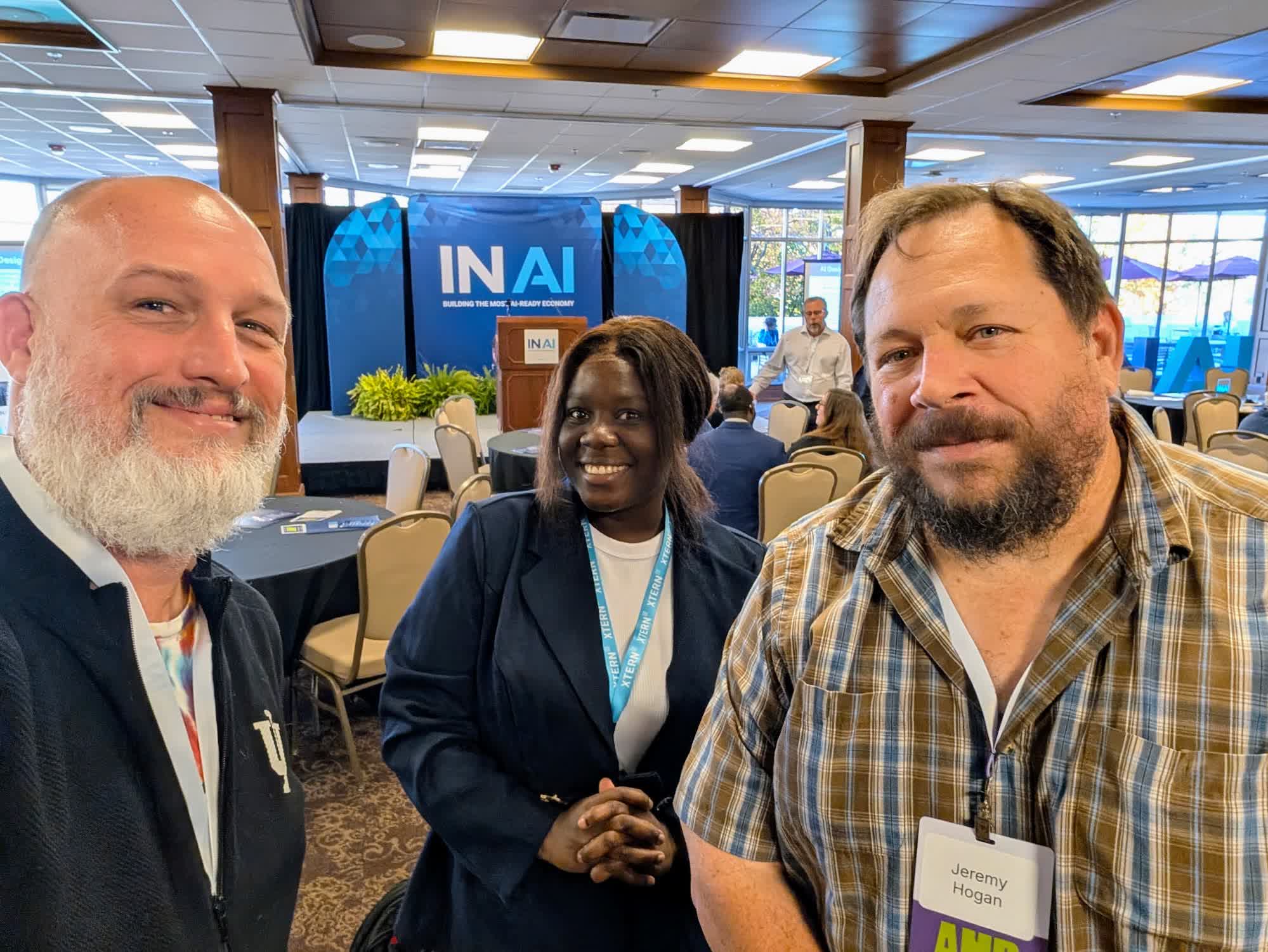 Three people pose for a selfie in the IN AI Roadshow room, with round tables and the IN AI podium behind them