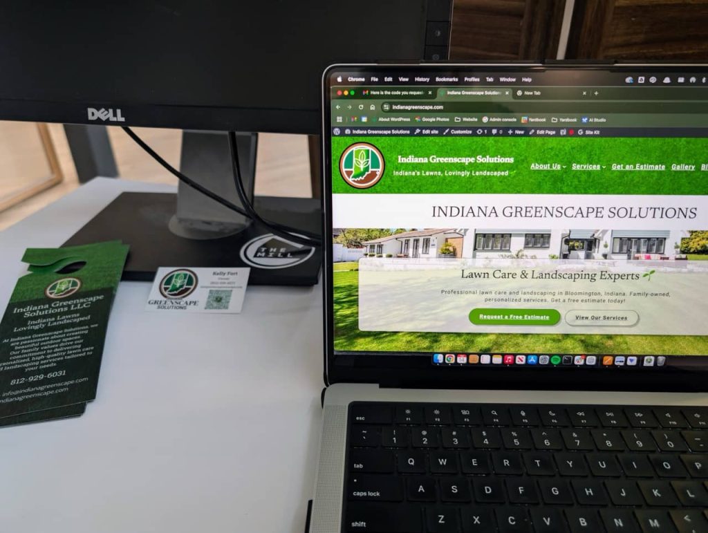 A workspace showing Indiana Greenscape Solutions branding materials, including a door hanger and business card, alongside a laptop displaying the company's website with a green-themed design.