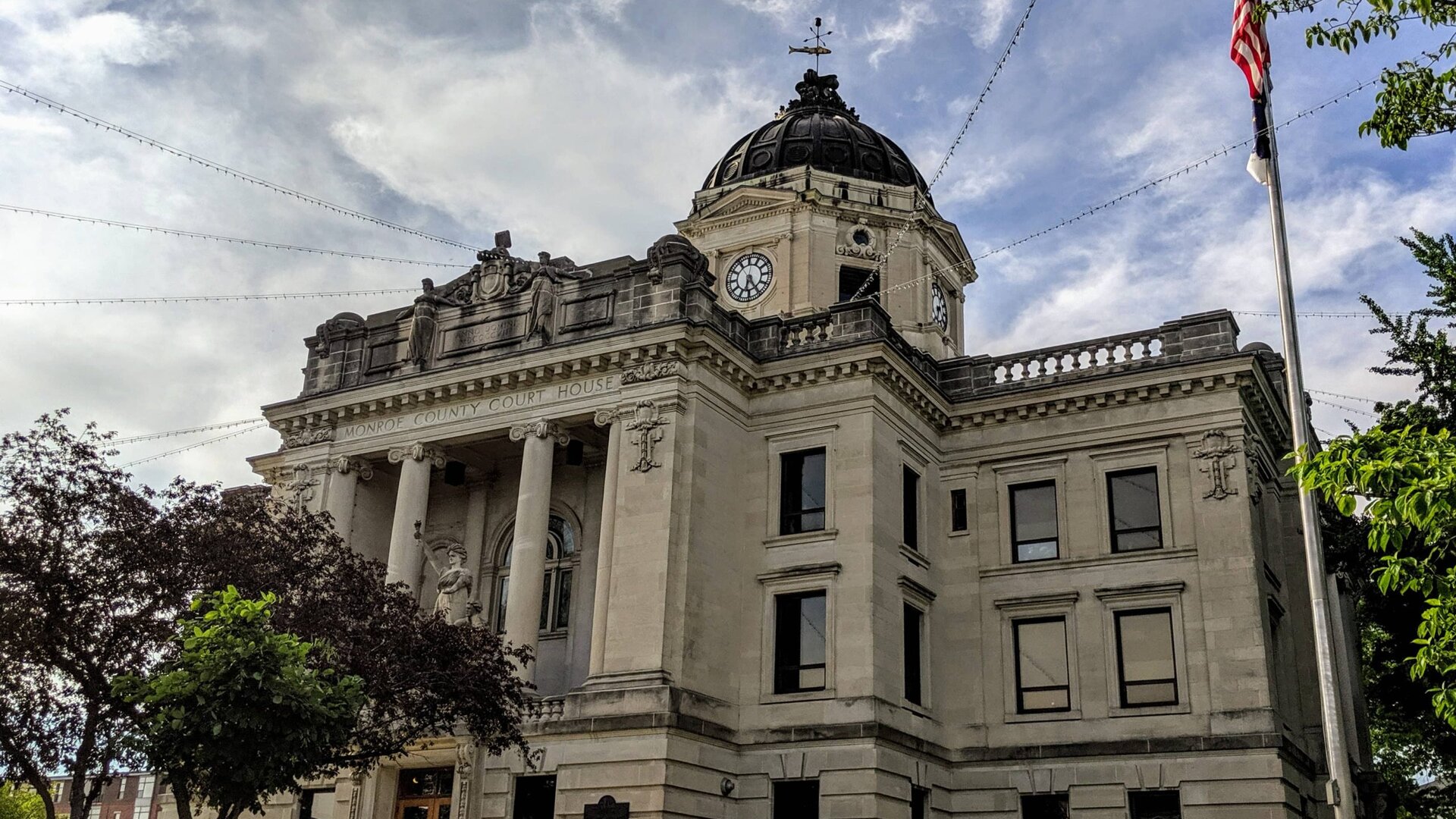 The Monroe County Courthouse in downtown Bloomington, Indiana, with its clock tower dome and an American flag against a cloudy sky.