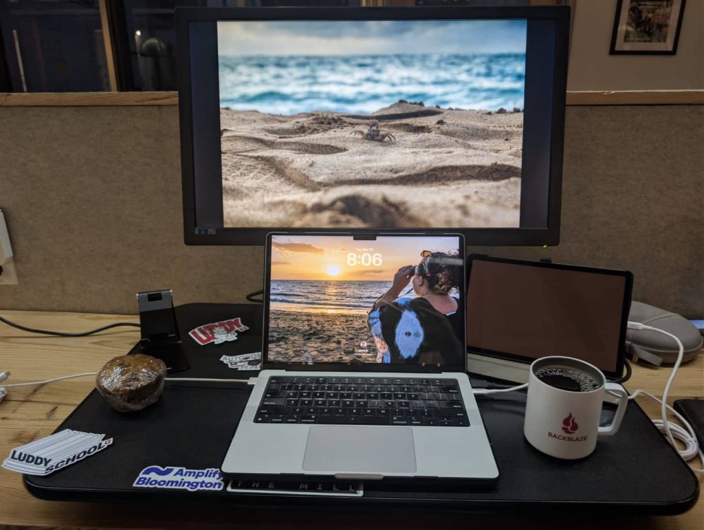 A multi-screen workspace at The Mill coworking space in Bloomington, Indiana, with a MacBook, external monitor, iPad, and a Backblaze coffee mug on a desk decorated with Amplify Bloomington stickers.