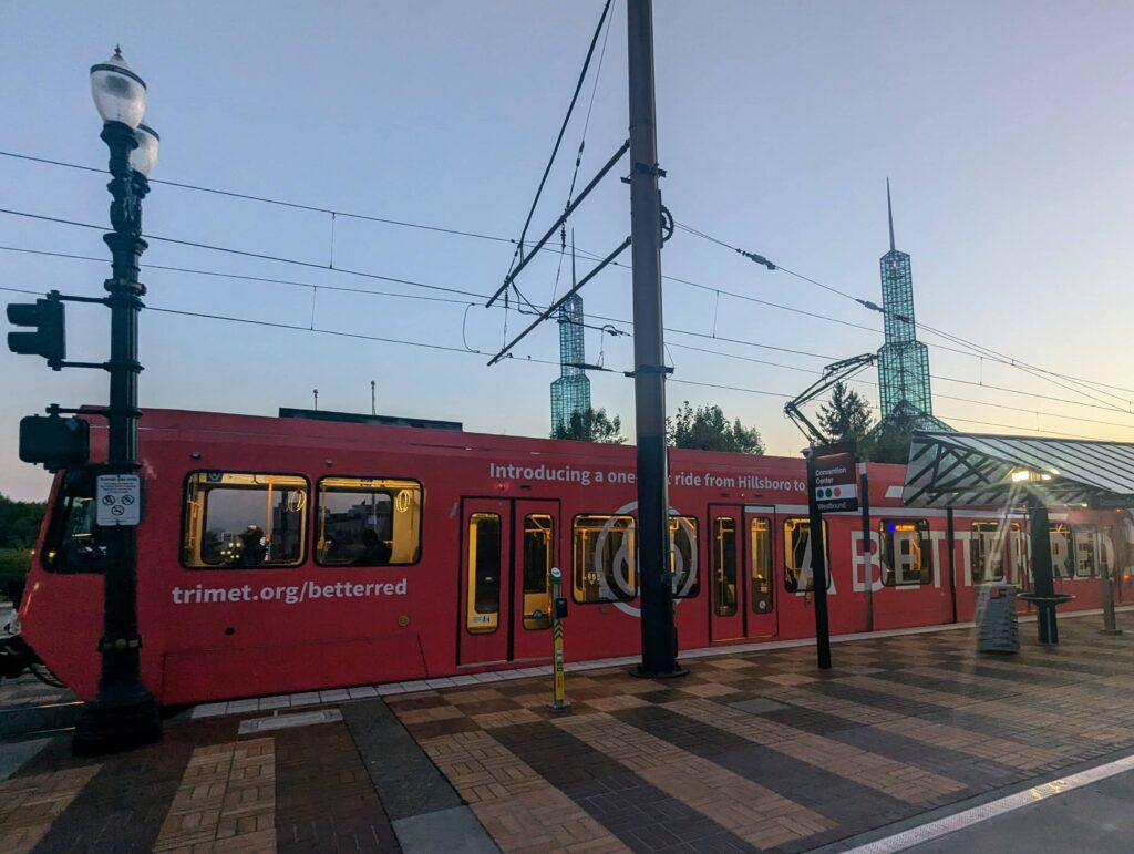 The Portland MAX Red Line train passing by the Oregon Convention Center at dusk, with the iconic twin glass spires in the background.