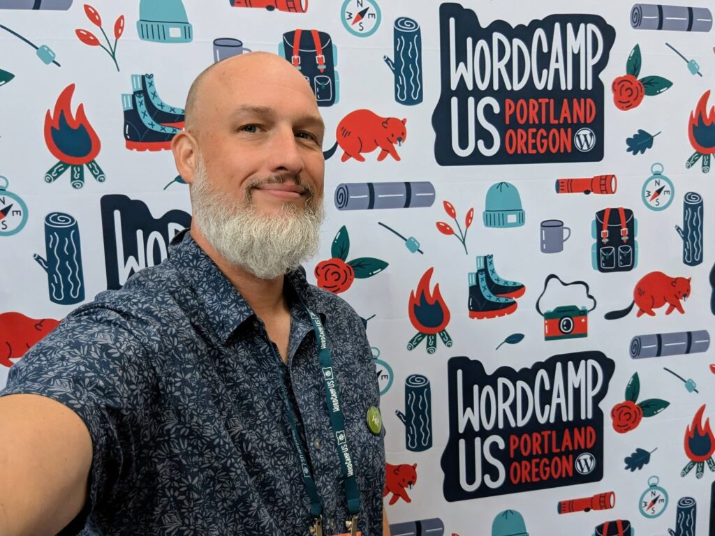 A man with a white beard, Dave Martin, takes a selfie in front of a WordCamp US 2024 backdrop in Portland, Oregon.