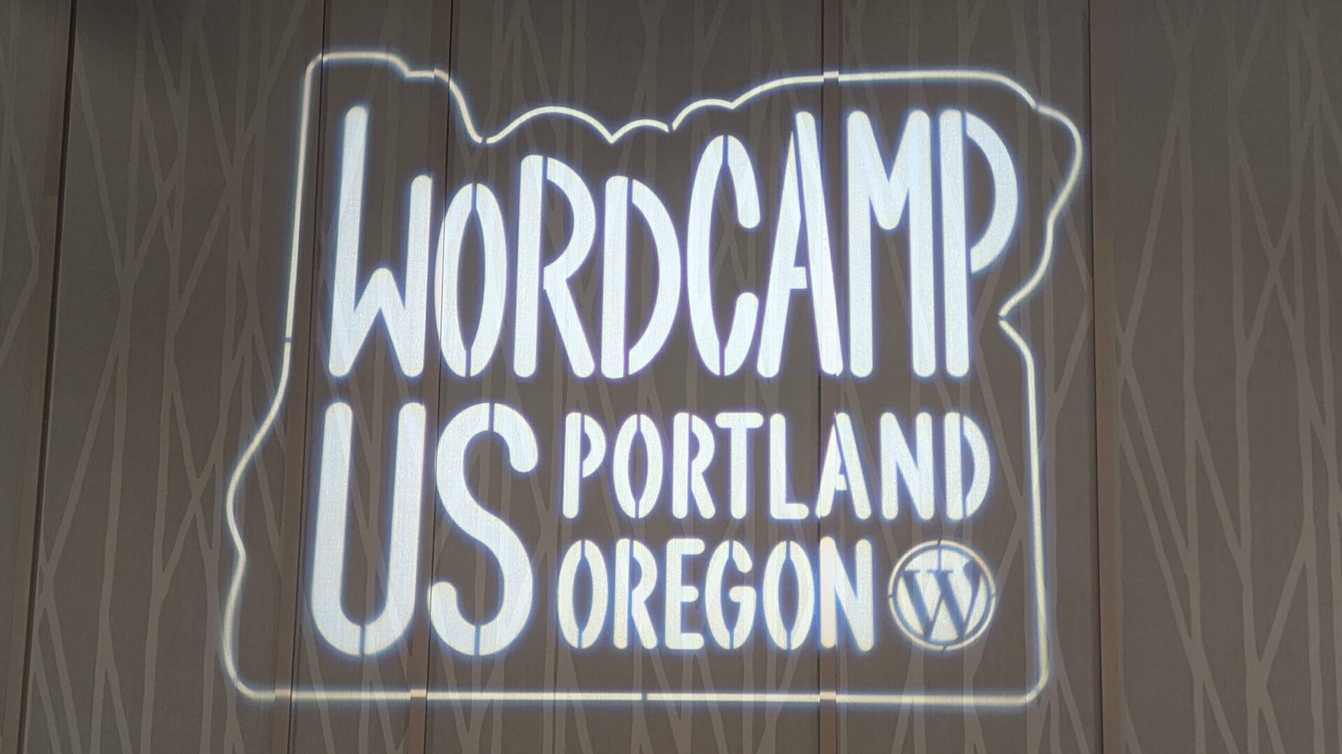The WordCamp US 2024 Portland, Oregon logo with the WordPress symbol, projected on a wall at the Oregon Convention Center.